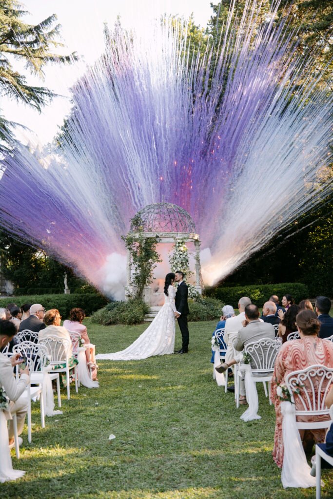 Wedding ceremony in a garden. The bride and groom are standing in front of an iron gazebo, while white and purple colored smoke bombs go off behind them.